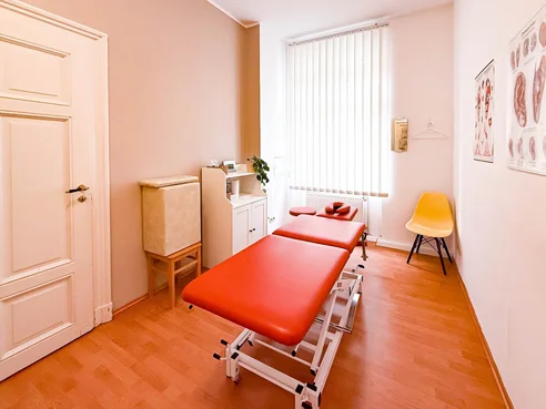 Behandlungsraum mit orangefarbener Therapieliege, gelbem Stuhl und Tageslicht.