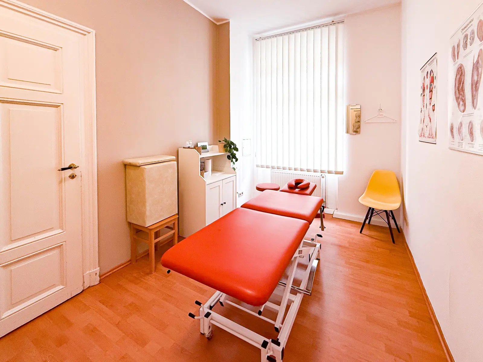 Behandlungsraum mit orangefarbener Therapieliege, gelbem Stuhl und Tageslicht.